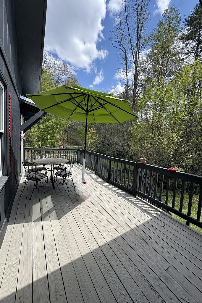 A wooden deck with a table and chairs under a green umbrella, surrounded by a railing and trees.