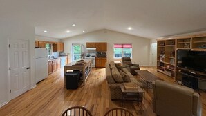 Amplia sala de estar con piso de madera, un sofá, un televisor y una cocina al fondo.