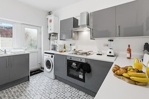 A modern kitchen with grey cabinets, a washing machine, and a breakfast bar with croissants and fruit. - WN1 Wigan Stay | 3 Bed | WiFi & Parking (Wigan)