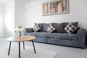 A living room with a grey sofa, a round wooden coffee table, and a white vase with flowers. - Haydock WA11 | Large 4 BR Home | Work & Group Stay (Haydock)