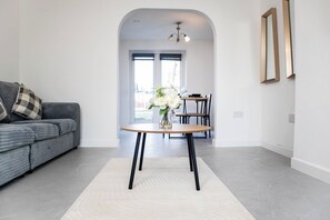 Ein modernes Wohnzimmer mit einem grauen Sofa, einem runden Holztisch mit schwarzen Beinen und einem weißen Teppich.