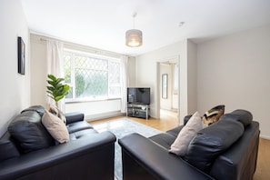 A modern living room with leather sofas, a flat-screen TV, a potted plant, and a large window with curtains. - Modern 3BR Home | Parking | Heart of West Didsbury (Manchester)