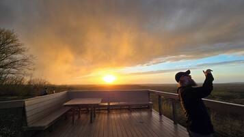 Eine Holzterrasse mit Bänken und einem Geländer, mit Blick auf eine Landschaft bei Sonnenuntergang.