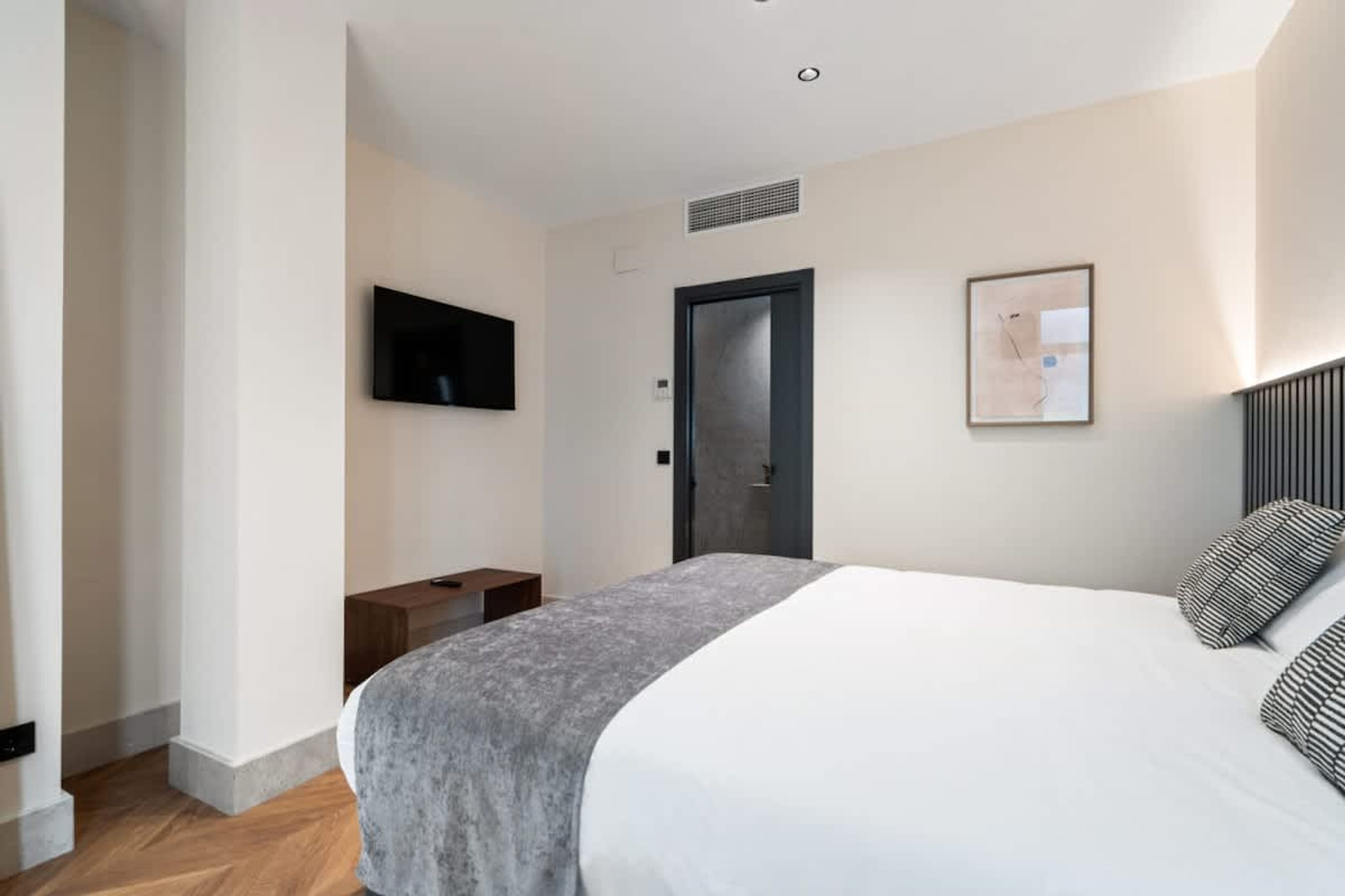 A modern hotel room with a large bed, a flat-screen TV mounted on the wall, a door with a handle, and a small wooden nightstand.