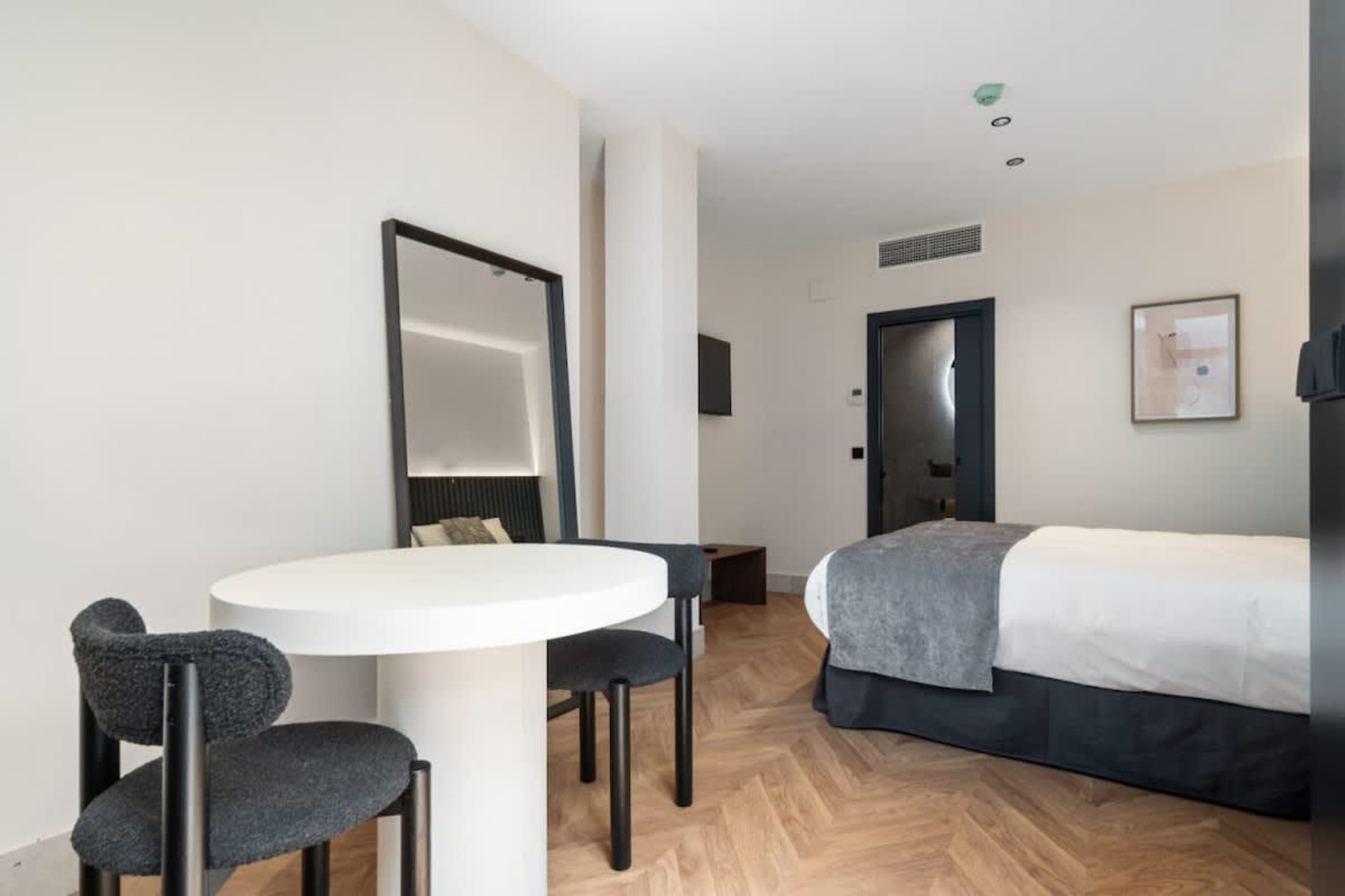 Una habitación de hotel moderna con una mesa redonda blanca, dos sillas, una cama con una colcha gris y un espejo grande en la pared.