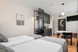Moderni hotellihuone, jossa on kaksi erillistä sänkyä, pieni keittonurkkaus, ruokailutila ja seinälle asennettu televisio.