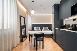 Moderni hotellihuone, jossa on pyöreä ruokapöytä, kaksi sänkyä, sisäänrakennettu keittiö ja suuri peili.