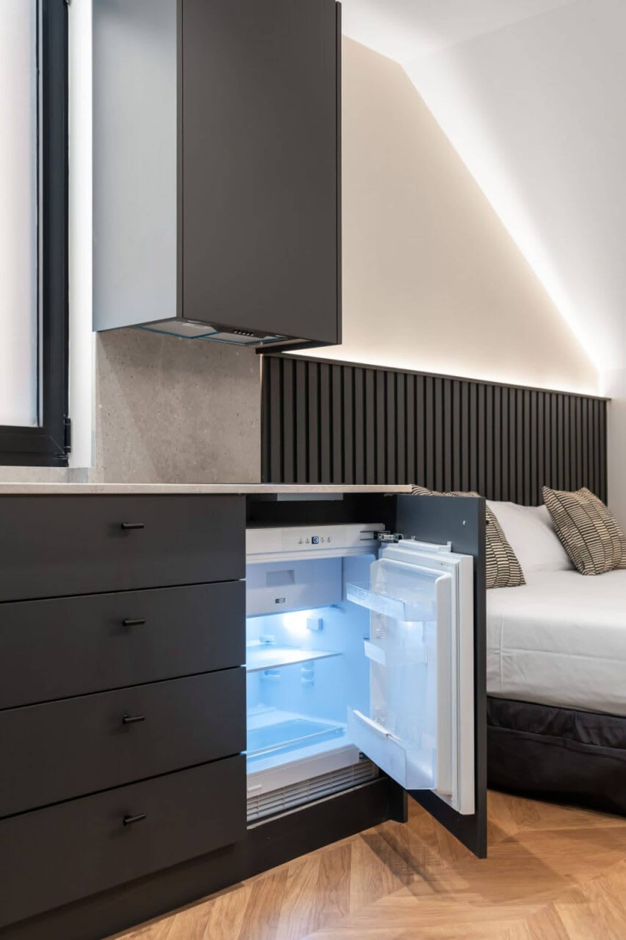 A modern kitchen with a built-in refrigerator, a dark wooden cabinet, and a bed with a striped pillow.
