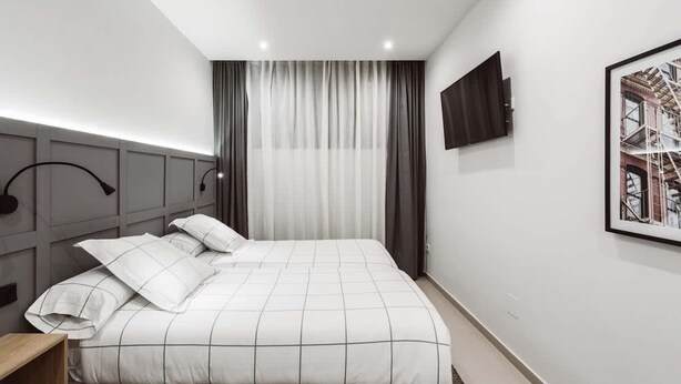 Una habitación de hotel moderna con una cama grande, un televisor de pantalla plana en la pared y una ventana con cortinas de gasa.