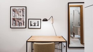 Una habitación con un escritorio, dos cuadros enmarcados, una lámpara y un espejo que refleja un baño.