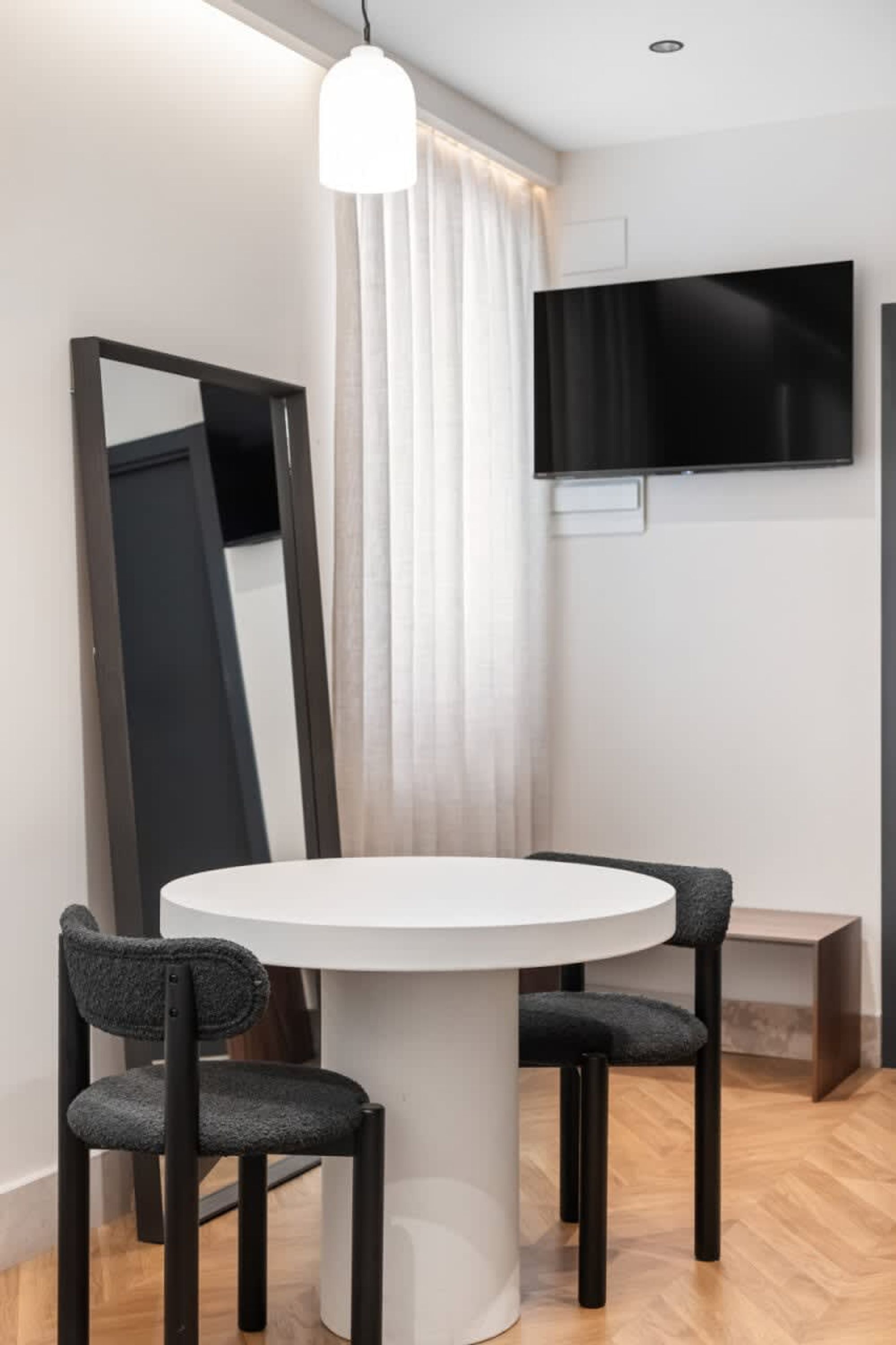 Una habitación moderna con una mesa de comedor redonda, dos sillas tapizadas, un espejo grande, un televisor de pantalla plana en la pared y una ventana con cortinas translúcidas.