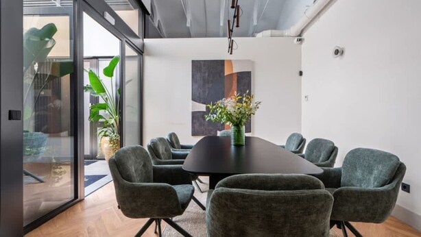 Een moderne vergaderruimte met een ronde tafel, grijs gestoffeerde stoelen en een vaas met bloemen.