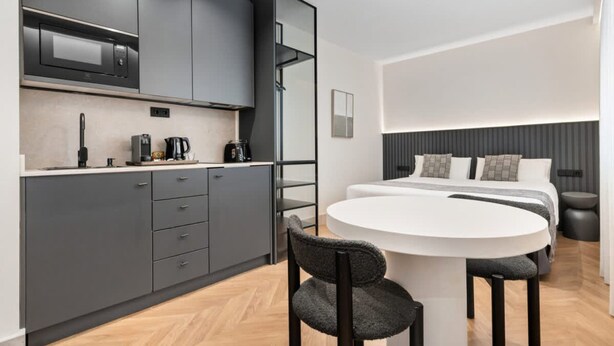 Una cocina moderna con armarios grises, microondas y cafetera. Hay una mesa de comedor redonda con dos sillas. La habitación también tiene una cama con cabecera gris y una mesita de noche.