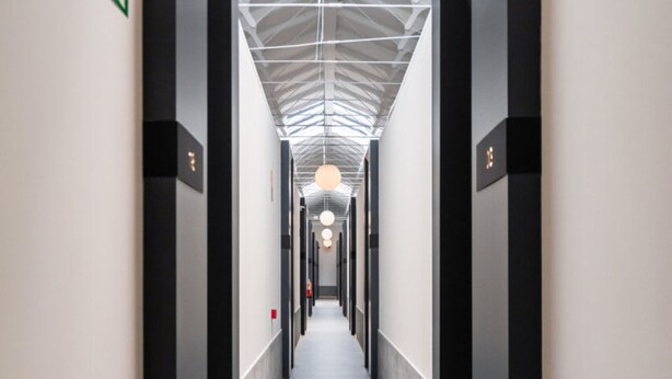 Un couloir d’hôtel moderne avec un plafond vitré, un panneau indiquant la sortie de secours et les portes d’un ascenseur.