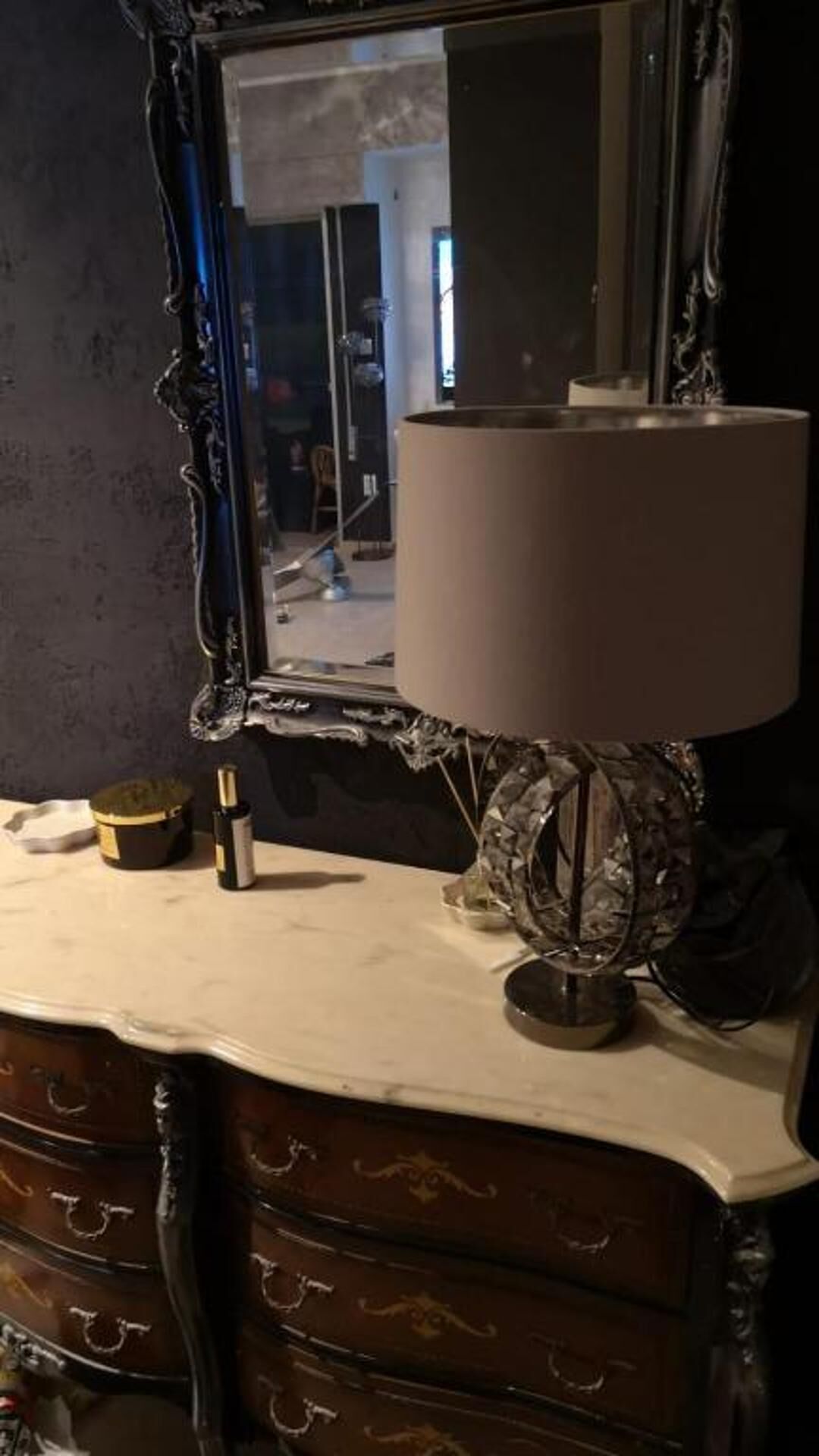A mirror with an ornate frame, a lamp with a white shade, and a small bottle on a dresser.