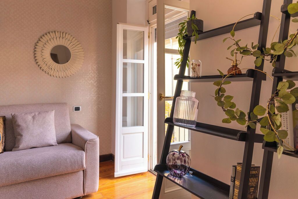 Ein Zimmer mit einer Couch, einem Zierteil-Spiegel, einem schwarzen Regal mit Pflanzen und einer Tür zu einem weiteren Raum.