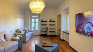 A living room with a chandelier, a sofa, an ottoman, and a television on the wall. - Beautiful spacious flat in the old town of Ivrea (Ivrea)