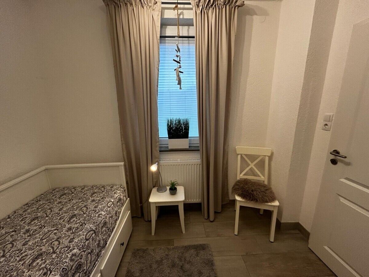 Een kleine, gezellige kamer met een bed, een stoel, een tafeltje en een raam met gordijnen.