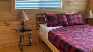 Puinen mökkimainen makuuhuone, jossa on sänky, kaksi yöpöytävalaisinta, ikkuna, jossa on sälekaihtimet, ja pieni pöytä, jossa on valaisin.