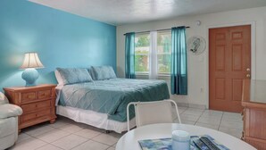 A bedroom with a bed, nightstand, chair, and a table with a lamp and remote controls. - Paradise Inn of Naples (Naples)