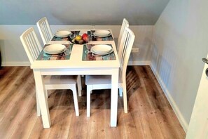 Une table à manger avec quatre chaises blanches, un panier de fruits et des verres à vin posés sur des sets de table.