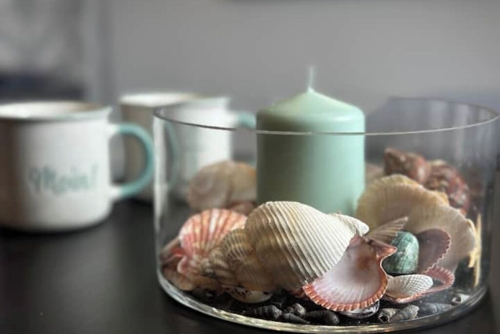 Een glazen houder met schelpen en een teal-kleurige kaars op een donker oppervlak.