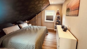 Un dormitorio con techo inclinado, una cama con edredón acolchado, una mesita de noche con lámpara y una ventana con persianas.