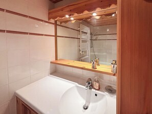 Ein Badezimmer mit einem weißen Waschbecken, einem Holzregal darüber und einem Spiegel, der einen Duschbereich reflektiert.