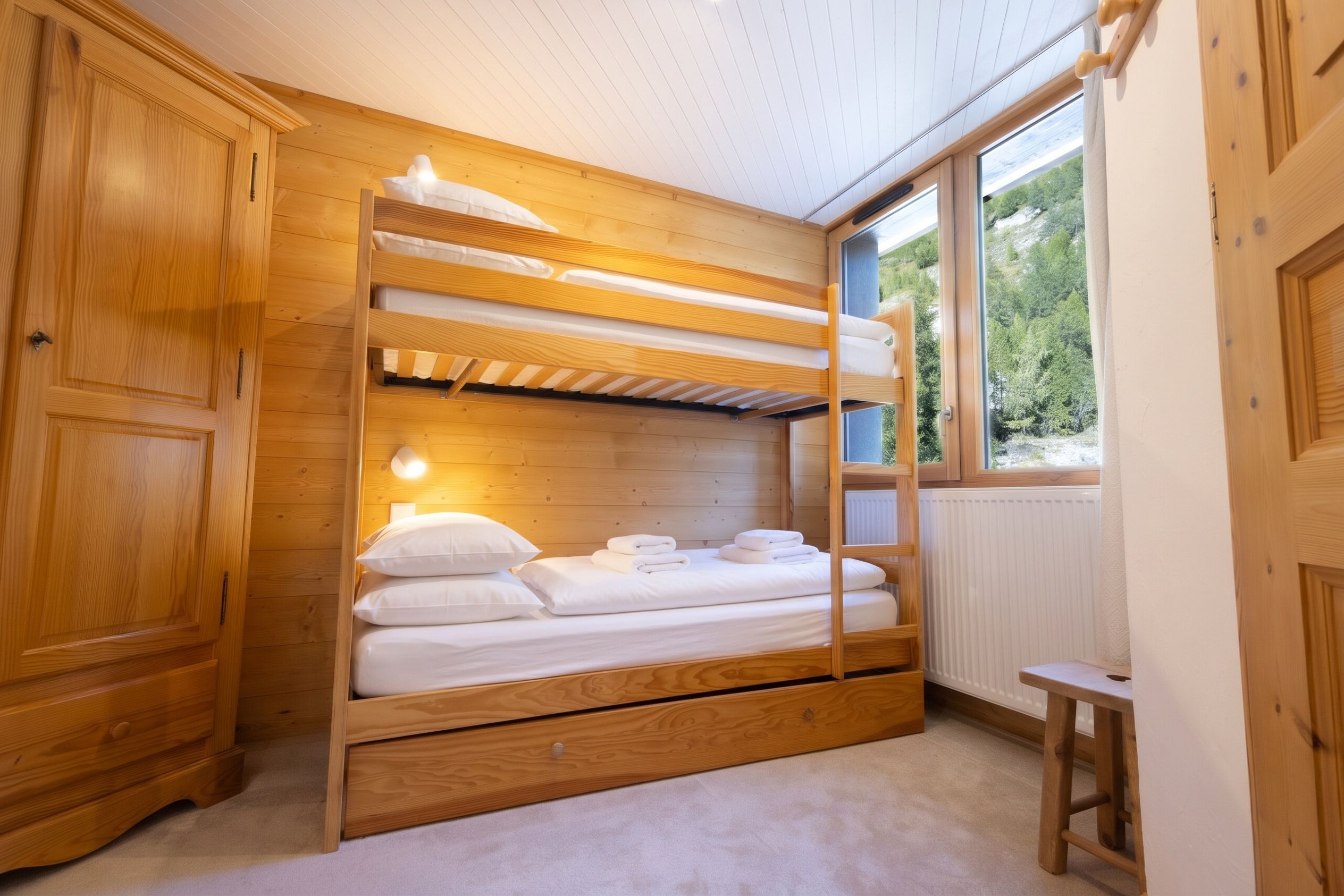 A bunk bed with a wooden headboard and a bedside table, a radiator, and a window with a view of greenery.