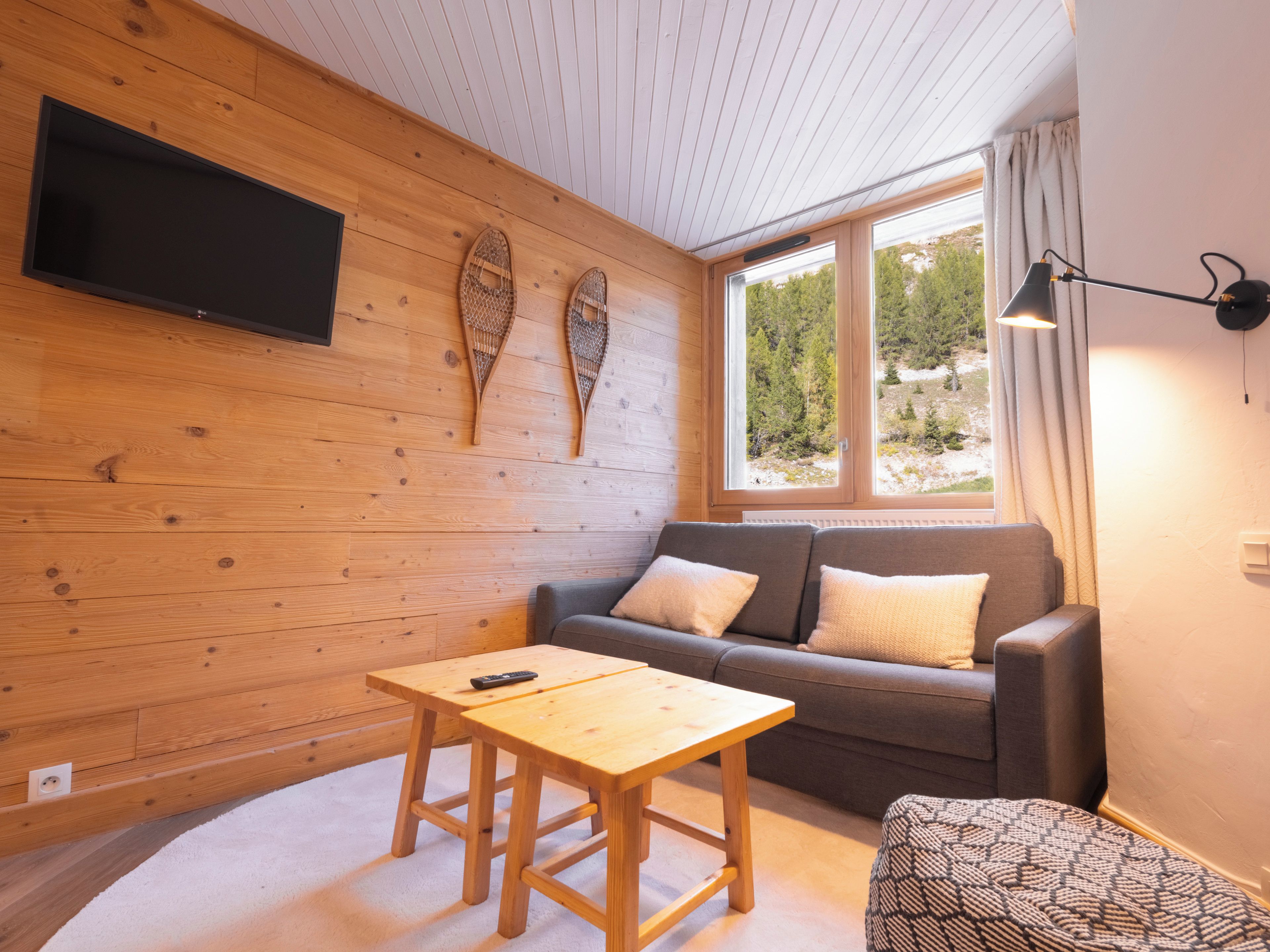 A cozy room with a wooden wall, a grey sofa, a small wooden table, and a window with a view of trees.