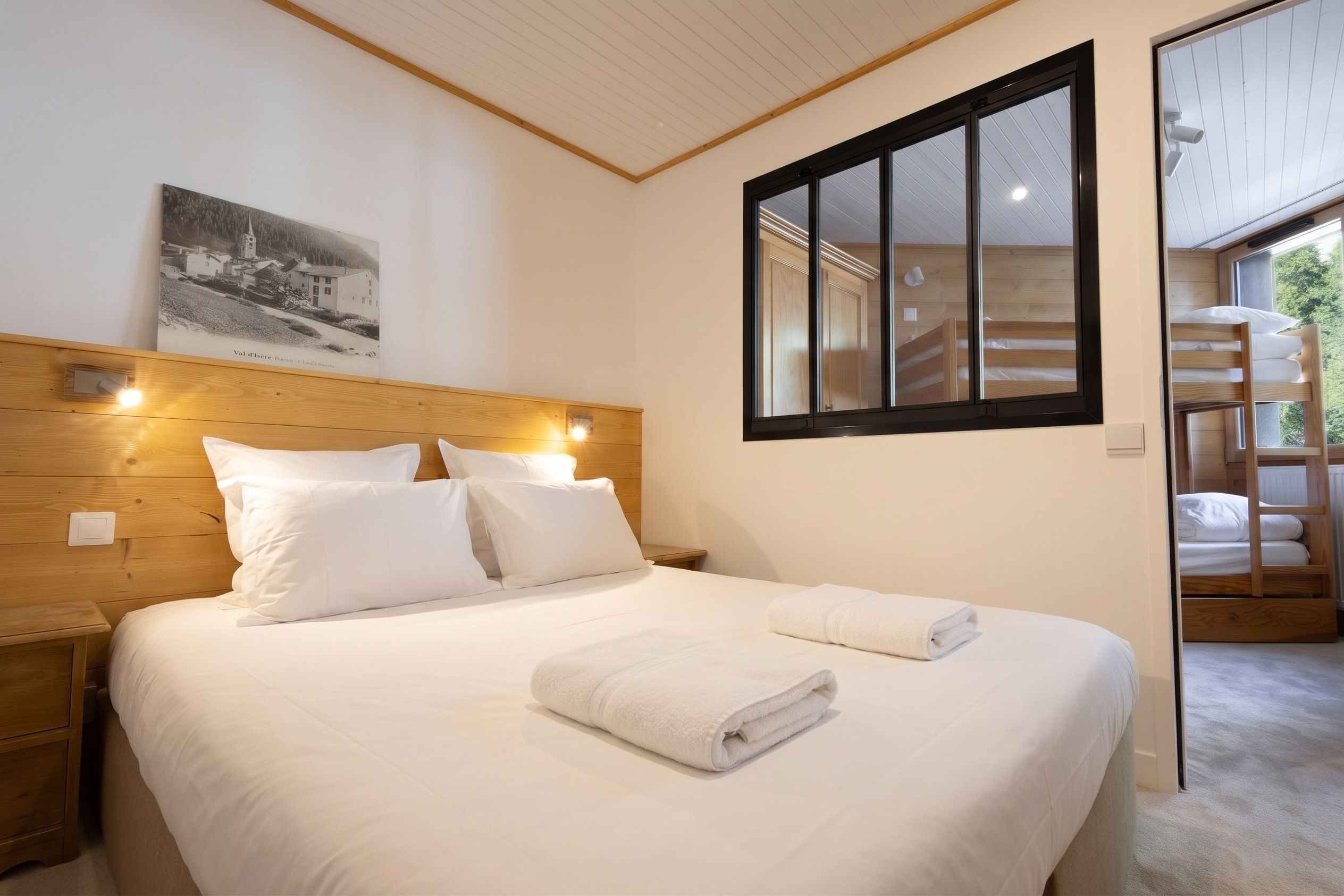 A bedroom with a wooden headboard, a bed with white linens, a rolled towel, and a window with a view of the outdoors.