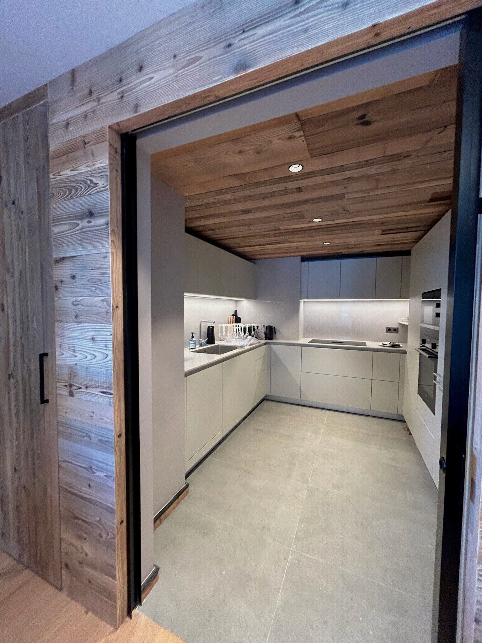 A modern kitchen with white cabinets, a wooden ceiling, and a tiled floor.