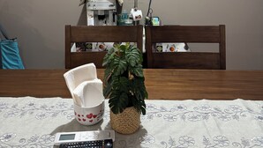 Uma área de refeições com uma mesa posta para dois, um controle remoto, uma caneca branca com corações vermelhos e um vaso de planta.