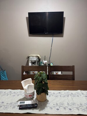 Een eethoek met een tafel gedekt voor twee personen, een afstandsbediening, een witte mok met rode harten en een bloempot met plant.