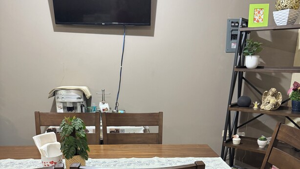 A dining area with a wooden table, chairs, a flat-screen TV mounted on the wall, a coffee machine, and a shelf with decorative items.