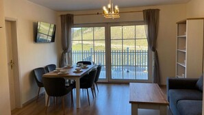 Een kamer met een eettafel voor vier personen, een bank, een aan de muur bevestigde televisie en een groot raam met uitzicht op een hek en groen.
