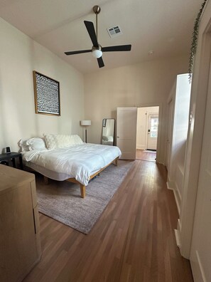 Ein Schlafzimmer mit Bett, Deckenventilator, Nachttisch, Lampe, Spiegel und einer Tür zu einem anderen Raum.