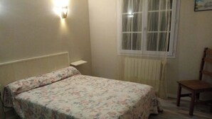 A bedroom with a floral bedspread, a wooden chair, a radiator, and a window with curtains.