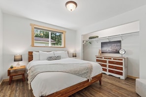 A bedroom with a wooden bed, a nightstand with a lamp, a dresser, a window, and a painting on the wall.