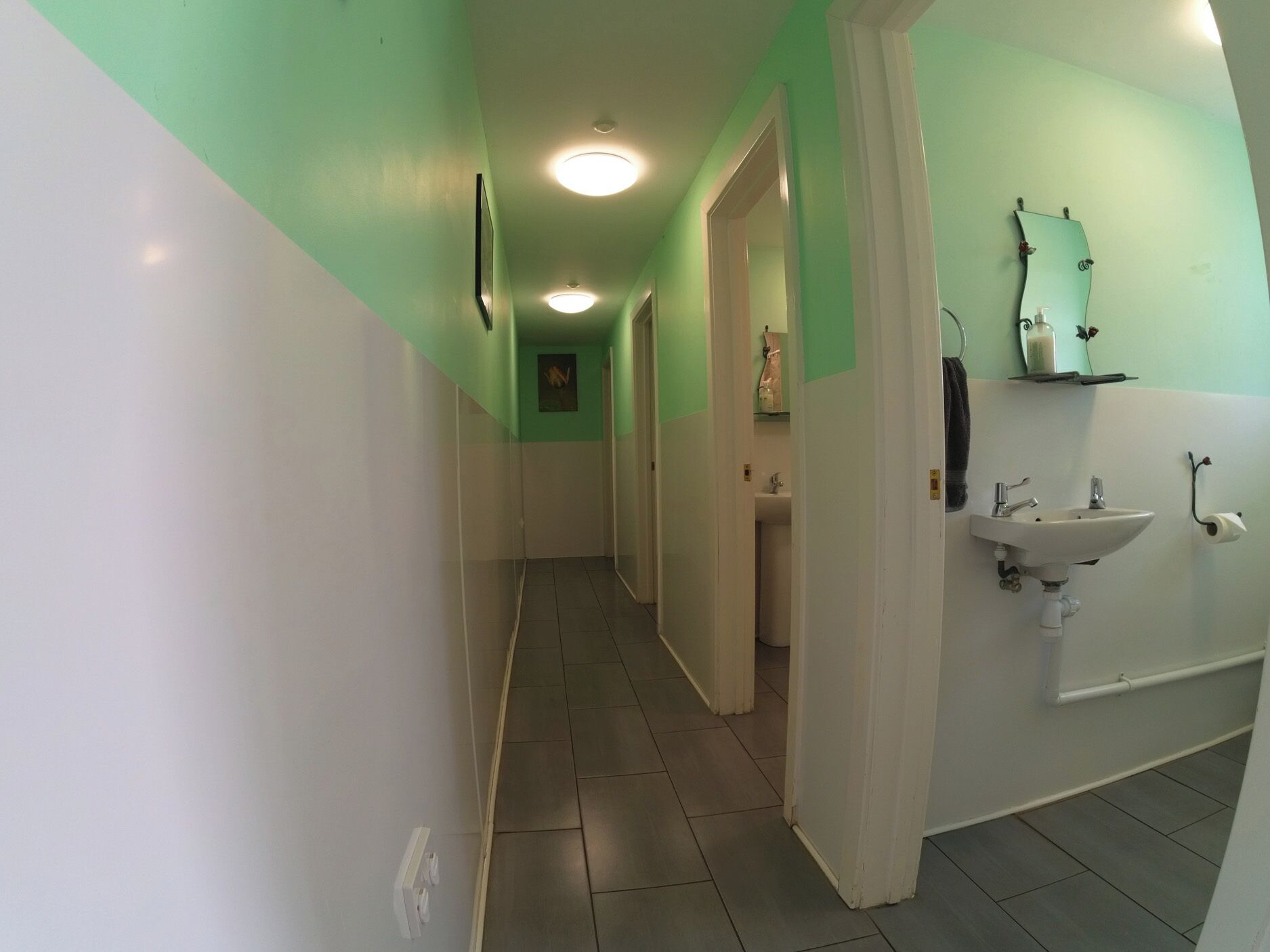 Een smalle gang met groene en witte wanden, een betegelde vloer, en een badkamer met een wastafel en spiegel aan de rechterkant.