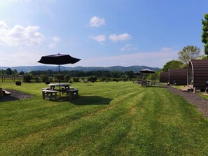 A well-maintained grassy area with picnic tables and umbrellas, surrounded by a wooden fence and a view of distant hills. - Blossom (Malvern)