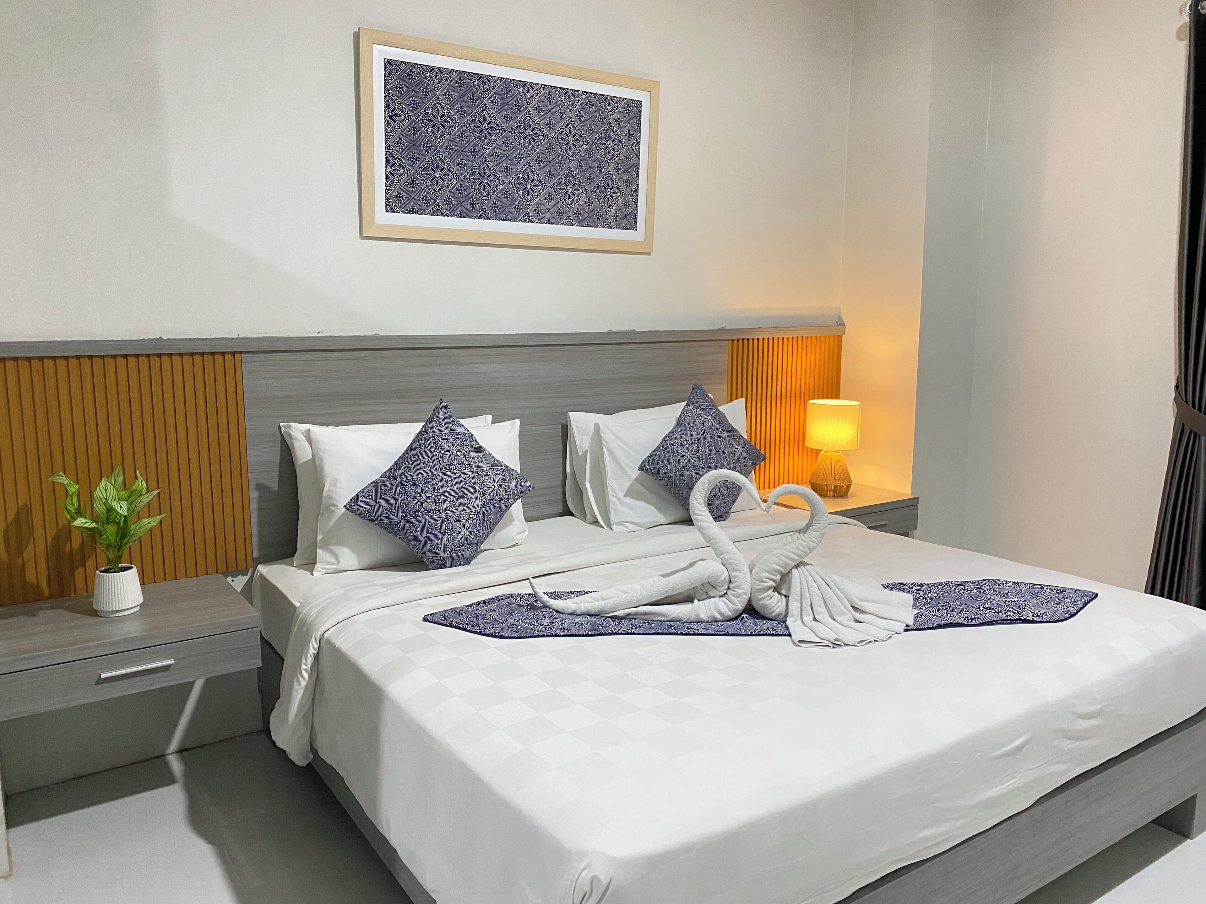 Una cama bien hecha con sábanas blancas y almohadones decorativos, una mesita de noche con una planta, una lámpara de pared y una obra de arte enmarcada sobre la cama.