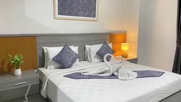 Una cama bien hecha con sábanas blancas y almohadones decorativos, una mesita de noche con una planta, una lámpara de pared y una obra de arte enmarcada sobre la cama.
