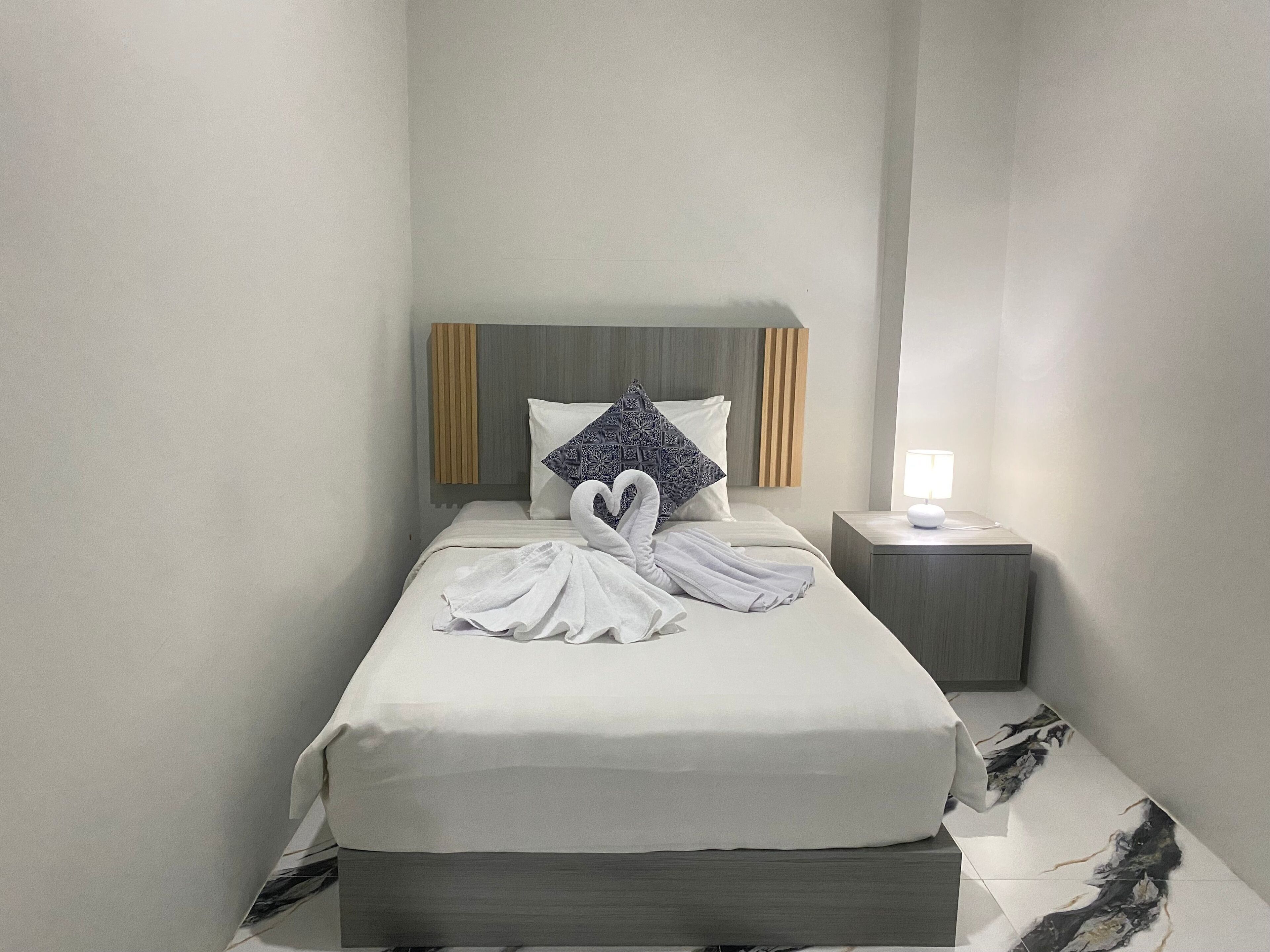 Een keurig opgemaakt bed met een handdoek in de vorm van een hart, een bedlampje en een nachtkastje met een lamp.