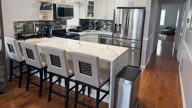 A modern kitchen with white cabinets, stainless steel appliances, and a marble island with bar stools. -  Spacious 3BR 2BA Apt Near JFK (Jamaica)