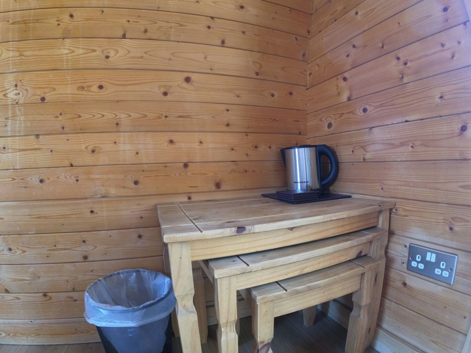 A wooden cabin interior with a wooden table, a stainless steel kettle, a trash bin, and a power outlet.