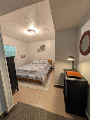 A bedroom with a bed, a bedside table with a lamp, a mirror, and a window with blinds. - Hummingbird Studio: 5 min to campus, quiet area (Starkville)