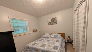 A bedroom with a bed, a window with blinds, a small table with a plant, and a picture on the wall.