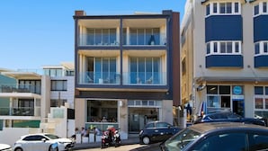 Un edifici modern de diverses plantes amb grans finestres i un balcó de vidre, situat en una zona urbana activa amb cotxes i vianants.