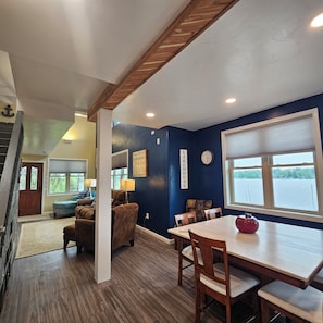 A dining area with a table and chairs, a clock on the wall, and a window with a view of a body of water. - Beautiful Lake Frontage house, handicap friendly, on Long Lake, Dundee Wisconsin (Campbellsport)